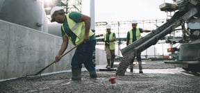 Construction crew pouring and leveling wet concrete on an industrial site, with a focus on one worker raking the freshly laid mix